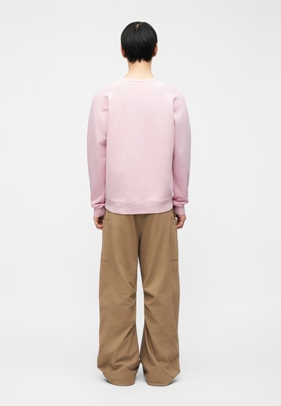 Personne aux cheveux courts noirs, vue de dos, portant un sweat-shirt rose clair et un pantalon marron ample, devant un fond blanc uni.