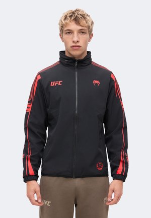 Un hombre rubio luce una chaqueta negra de UFC con cremallera, franjas rojas y logotipos, combinada con unos pantalones marrones sobre un fondo claro y liso.