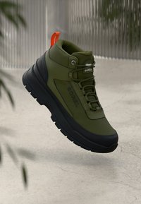 Olivengrøn høyhælte sneakers med svart såle, som har en jevn tekstur, rund form og oransje trekkløkke; logo synlig på siden.