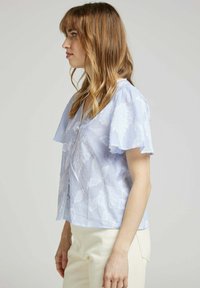 Blouse rayée bleue avec un motif floral, manches courtes volantées, décolleté en V et fermeture à boutons à l'avant. Texture de tissu douce.