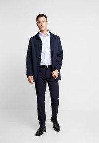 Marineblauw jasje met knopen en een kraag, gedragen over een lichtblauw overhemd en donkere broek. Zwarte schoenen maken de outfit compleet. Minimalistisch ontwerp.