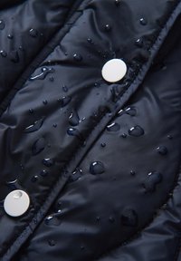 Tessuto imbottito impermeabile di colore blu navy con gocce d'acqua visibili; presenta bottoni a pressione bianchi e un design trapuntato, evidenziando la texture e il materiale.