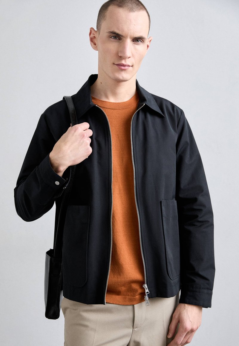 Sort lynlåsfutteral med spidse krave, frontlommer, båret over en orange sweater. Modellen holder en sort skuldertaske og har lysfarvede bukser på.