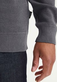 Pelēks struktūrēts džemperis ar ribotu apakšējo malu un piedurknēm, ar sānu birku. Roka atpūšas blakus tumši zilo audumu, piedāvājot tuvplāna skatu.