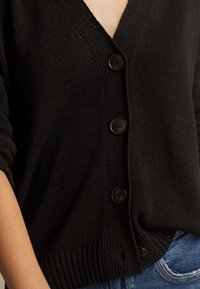 Schwarze Strickjacke mit V-Ausschnitt, fünf Knöpfen, geripptem Saum und glatter Textur. Details umfassen mittelgroße, dunkle Knöpfe.