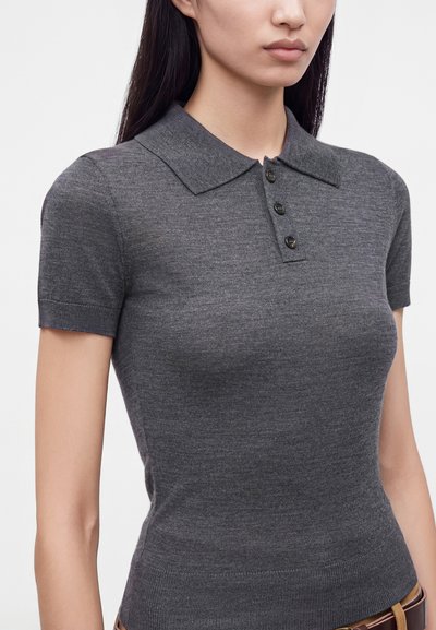 Femme portant un polo gris ajusté à manches courtes avec trois boutons, debout devant un fond blanc uni.