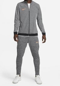 Gray Nike tracksuit with zippered jacket and tapered pants. Features orange logo accents and ribbed cuffs. Worn with white athletic shoes.