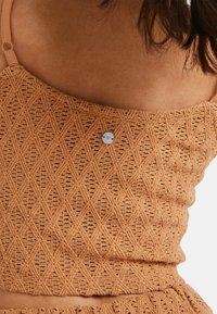 Woman wearing a textured, diamond-patterned, tan knit spaghetti strap top with a small round metal brand tag at the center chest.