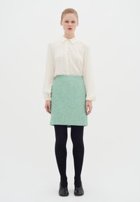 Cremefarvet bluse med knapper, grøn mønstret nederdel, sorte strømpebukser og sorte sko. Glat stof og skræddersyet pasform. Enkel krave design.