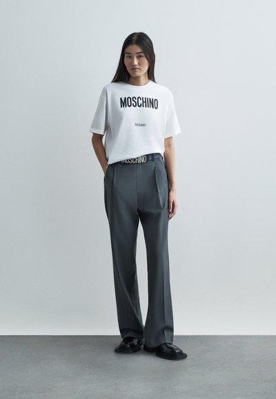 Mladá žena stojící s dlaněmi v kapsách, oblečená v bílem tričku Moschino Milano, šedých plisovaných kalhotách a černých botách, na neutrálním šedém pozadí.