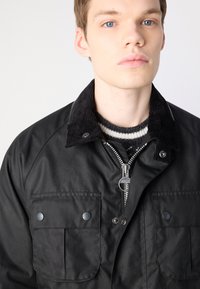 Black jacket with a corduroy collar, zip closure, and two front pockets. Features a smooth texture and silver zip hardware.