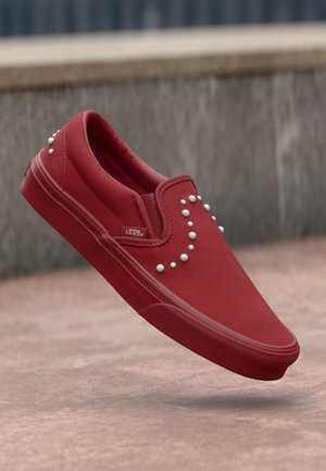 Piros vászon slip-on sportcipő kerek fehér szegecsekkel, sima textúrával, lekerekített orral és gumi talppal. Márkás logó az oldalon.