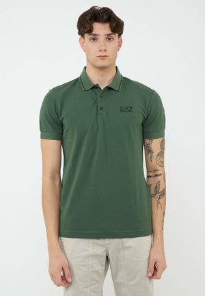 Giovane uomo che indossa una polo verde EA7 e pantaloni beige chiaro, in piedi con un'espressione neutra e tatuaggi visibili sul braccio sinistro.