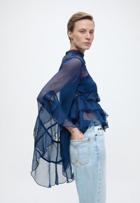 Γυναίκα με διάφανη μπλούζα σε σκούρο μπλε χρώμα με βολάν και ανοιχτόχρωμα τζιν, στέκεται πλάγια με το χέρι στην πίσω τσέπη μπροστά σε λευκό φόντο.