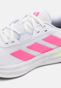 Baskets blanches de sport avec un dessus en mesh, trois bandes roses proéminentes sur le côté et des détails texturés. Elles sont dotées de lacets blancs et d'une semelle rembourrée.
