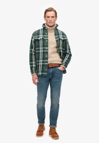 Camisa de franela a cuadros en verde y blanco, cuello alto beige, jeans azules y botas de cuero marrón. Incluye un cinturón marrón y un reloj.