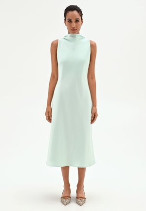 Donna in piedi rivolta in avanti che indossa un abito midi verde chiaro senza maniche e décolleté beige con punta appuntita su sfondo bianco uniforme.