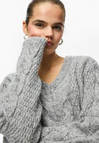 Jeune femme aux cheveux plaqués en arrière, portant des boucles d'oreilles argentées et un gros pull en maille torsadée gris, avec la main partiellement couvrant sa bouche.