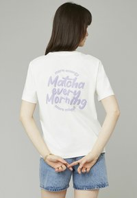 T-shirt en coton blanc avec un texte graphique violet au dos portant "plus d'énergie Matcha chaque matin, plus conscient", associé à un short en jean bleu.