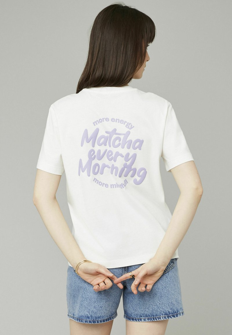 T-shirt en coton blanc avec un texte graphique violet au dos portant "plus d'énergie Matcha chaque matin, plus conscient", associé à un short en jean bleu.
