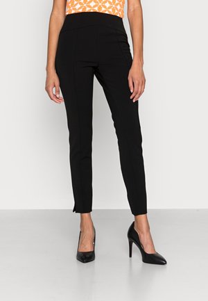Pantalones negros ajustados hasta el tobillo llevados con tacones altos negros y una blusa sin mangas naranja con estampado, de pie frente a un fondo liso.