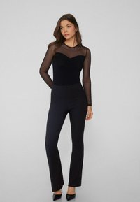 Černý overal s dlouhými průhlednými rukávy, přiléhavým tělem a rozšířenou nohavicovou verzí. Hladký materiál s detailem ve tvaru srdce u výstřihu.