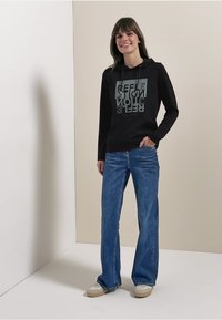 Schwarzer Hoodie mit grafischem Textdesign, kombiniert mit hellblauen Schlagjeans und beigen Sneakern. Glatter Stoff, lässige Passform, Basisfarben.