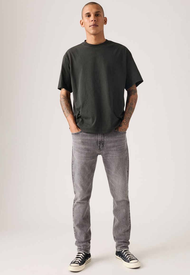 Camiseta de cuello redondo verde oscuro, corte relajado, combinada con jeans gris claro y zapatillas negras. Diseño sencillo sin patrones ni logotipos visibles.