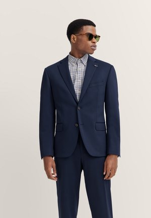Mann trägt marineblauen Anzug mit geschlossenem Blazer, kariertem Hemd und Sonnenbrille, schaut nach rechts vor einfarbigem Hintergrund.