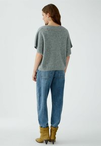 Frau in einem lockeren grauen kurzärmligen Pullover, blauen Jeans und beigen Wildleder-Stiefeln mit Absatz, die mit dem Rücken zu einer einfarbigen Hintergrund steht.