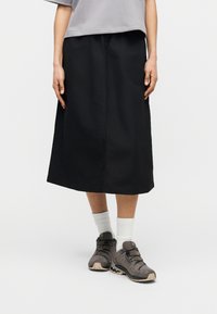 Άτομο που φορά μαύρη φούστα μέχρι το γόνατο, γκρι πουκάμισο, λευκές κάλτσες και γκρι παπούτσια πεζοπορίας, στέκεται σε απλό φόντο.