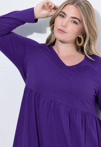 Langes lila Oberteil mit V-Ausschnitt und ausgestelltem Saum. Weicher Stoff mit gerafften Details, kombiniert mit großen strukturierten Creolen.