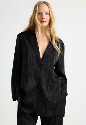 Nainen, jolla on pitkät vaaleat hiukset ja yllään suuri musta pinstripe-bleiseri, jossa on kaksi nappia, ja yhteen sopivat housut, kädet taskuissa, neutraali tausta.