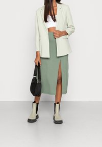 Světle zelené sako přes bílé cropped tričko, doplněné sukní do kolen zelené barvy s předním rozparkem. Černé kotníkové boty a černá taška.