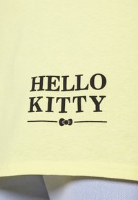 Camiseta de algodón amarilla con "HELLO KITTY" impreso en letras grandes negras y acentos decorativos debajo del texto.
