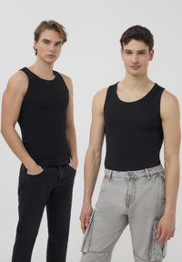 Due modelli maschili che indossano canotte nere aderenti. Un paio di jeans scuri, un paio di pantaloni cargo grigio chiaro con tasche laterali. Sfondo semplice.