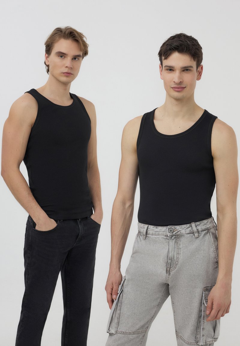 Due modelli maschili che indossano canotte nere aderenti. Un paio di jeans scuri, un paio di pantaloni cargo grigio chiaro con tasche laterali. Sfondo semplice.