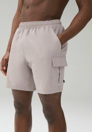 Męski tors i nogi w beżowych szortach cargo z boczną kieszenią, jedna ręka w kieszeni, na jasnym jednolitym tle.