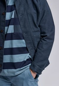 Veste bleu marine avec fermeture à boutons et poches latérales, superposée à une chemise à rayures bleues et marine. Tissu doux, coupe décontractée.