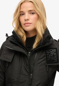 Veste noire isolante avec un col montant, deux poches zippées, surface texturée et un badge avec "SuperDry" sur l'épaule.