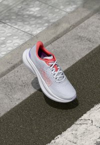 Chaussure de course gris clair avec une tige en mesh texturé, des accents roses au niveau du collier et du logo, et une semelle blanche amortissante.