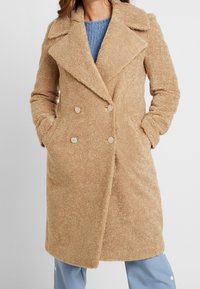 Cappotto doppiopetto beige realizzato in tessuto testurizzato con colletto, dotato di bottoni dorati e tasche laterali. Indossato sopra un maglione blu.