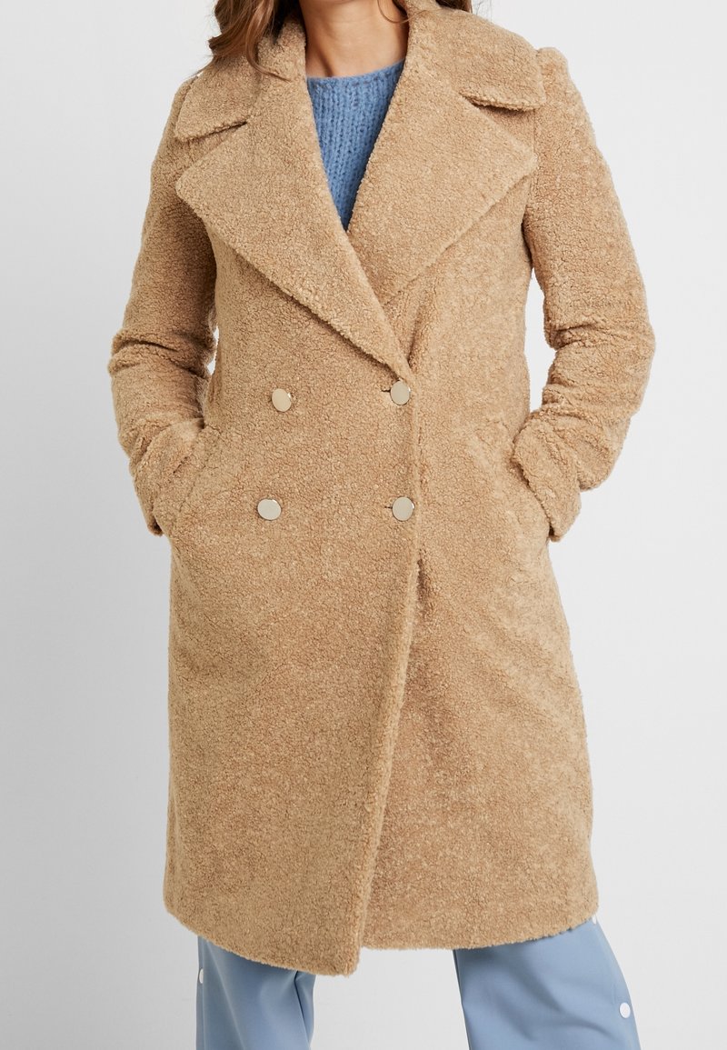 Cappotto doppiopetto beige realizzato in tessuto testurizzato con colletto, dotato di bottoni dorati e tasche laterali. Indossato sopra un maglione blu.
