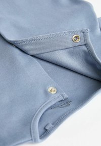 Světle modrá látka s hladkým povrchem, se zapínáním na patent a žebrovanými okraji. Design zahrnuje štítek a jemný detail švu.
