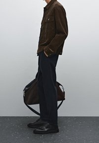 Brązowa kurtka sztruksowa z dwoma kieszeniami na piersi, zestawiona z ciemnymi spodniami i czarnymi sznurowanymi butami. Przez ramię przewieszona jest brązowa torba typu tote.