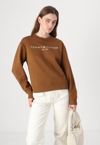 Hnědá mikina s kulatým výstřihem a bílým nápisem "TOMMY HILFIGER EST. 1985". S žebrovanými manžetami a lemem, nošena s bílými kalhotami a béžovou taškou.