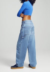 Osoba ubrana w niebieski krótki top z długim rękawem i luźne jasnoniebieskie dżinsy, w granatowych trampkach, stojąca na tle jednolitego jasnego tła.