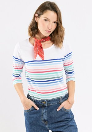 Femme portant un t-shirt à manches longues à rayures horizontales multicolores, un jean bleu et un foulard rouge à motifs autour du cou, les mains dans les poches.