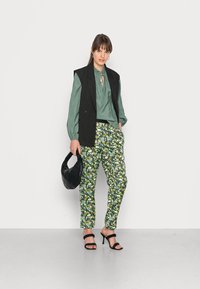 Blusa de cetim verde com decote franzido, colete risca de giz preto, calças tapered com estampa floral, bolsa acolchoada preta e sapatos de salto preto com tiras.