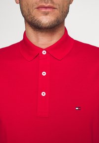 Polo rouge avec un col classique, quatre boutons de fermeture avec des coutures rouges contrastantes, et un petit logo brodé sur la poitrine gauche.